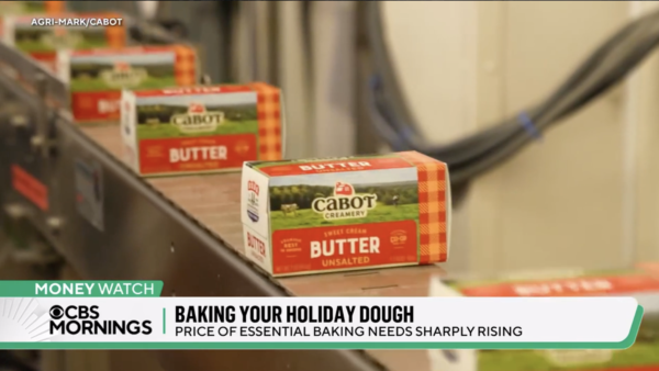 Boxes of unsalted butter move along a conveyor belt. A news headline reads: "Baking Your Holiday Dough: Price of Essential Baking Needs Sharply Rising.
